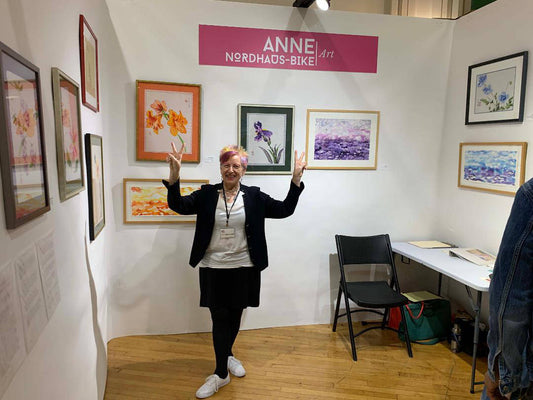 Anne Nordhaus-Bike posing in exhibit booth in front of her framed watercolors with logo sign above.