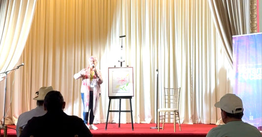 Artist standing on a stage with framed watercolor painting, large curtain backdrop, people in foreground.