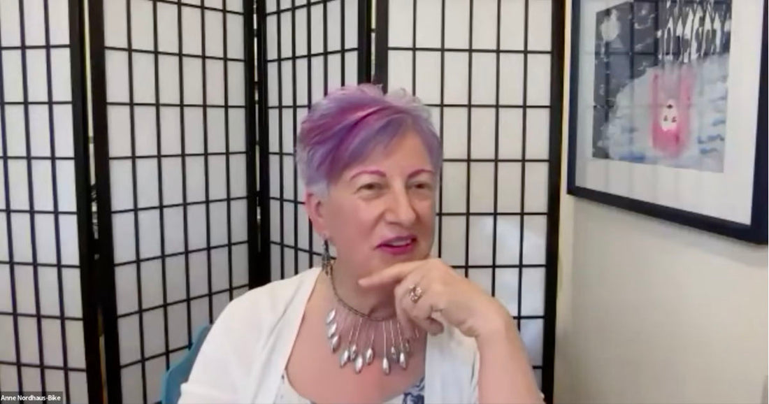 Woman with short purple hair wearing a white top and a silver statement necklace, gesturing with her hand while speaking in front of a black-and-white screen.