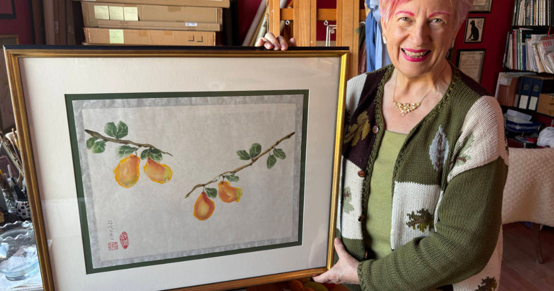 Smiling woman in art studio holding large framed watercolor of yellow pears on branches with green leaves.