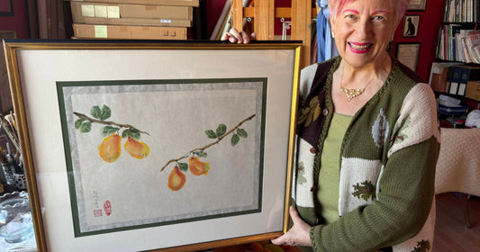 Smiling woman in art studio holding large framed watercolor of yellow pears on branches with green leaves.