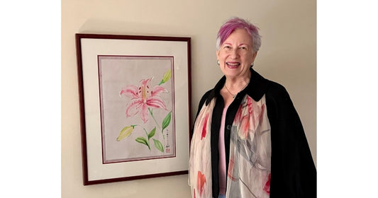 Smiling artist standing next to a framed watercolor painting of flowers on a wall.