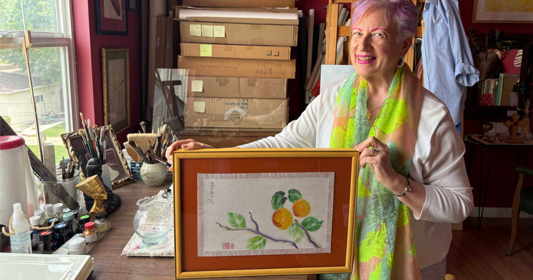 Woman in an art studio holding a framed watercolor of peaches on a branch.
