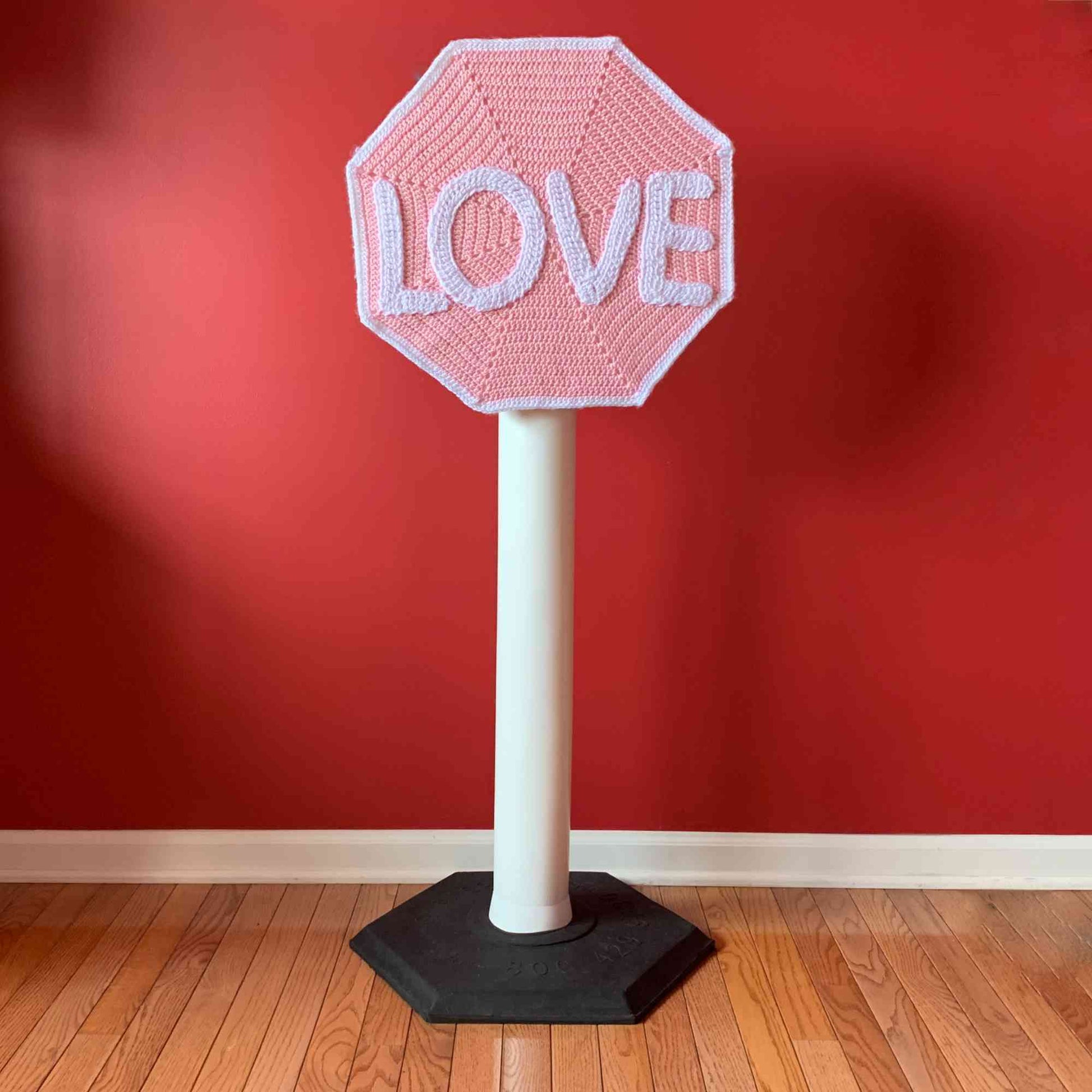 Love Sign pink and white crocheted stop sign mixed media art, full view in front of red wall