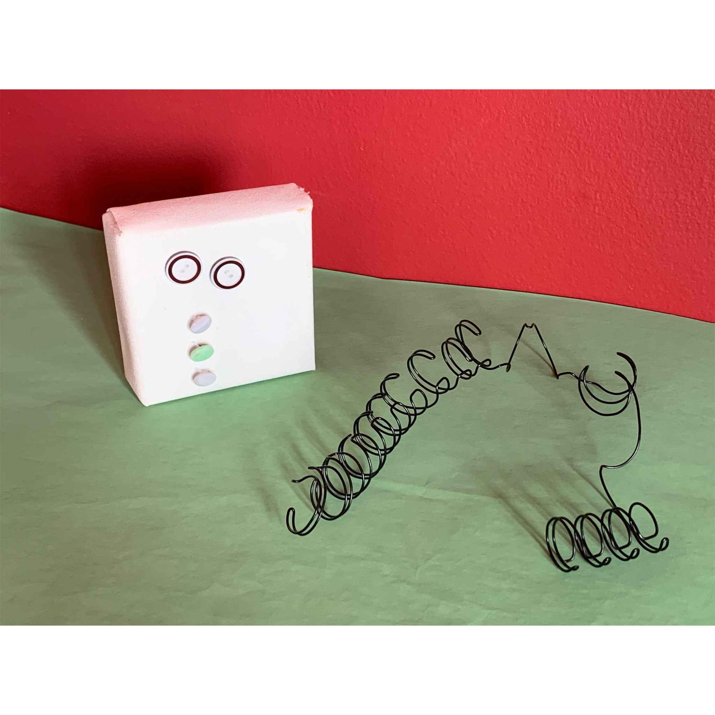 Wirehaired Me multimedia sculpture on green surface with red wall, hair sitting next to "face"